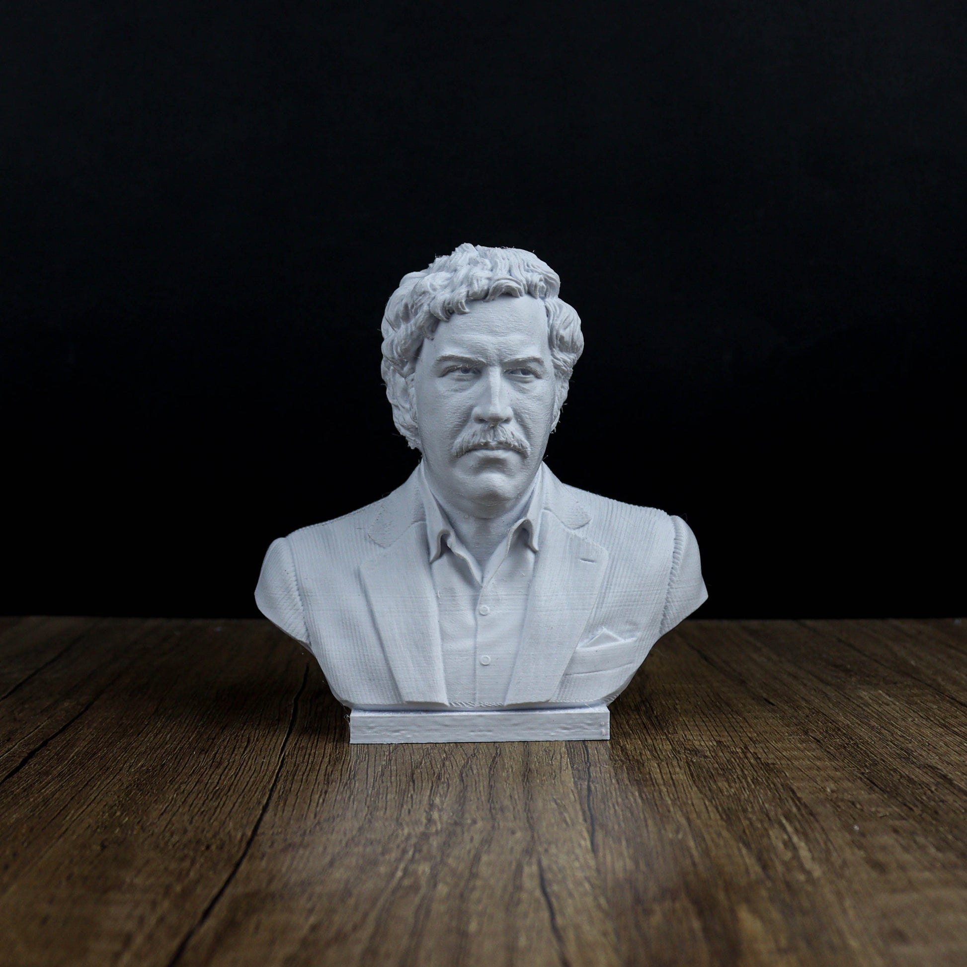 A white bust of a man with curly hair and a mustache, wearing a suit, is displayed on a wooden surface against a dark background.