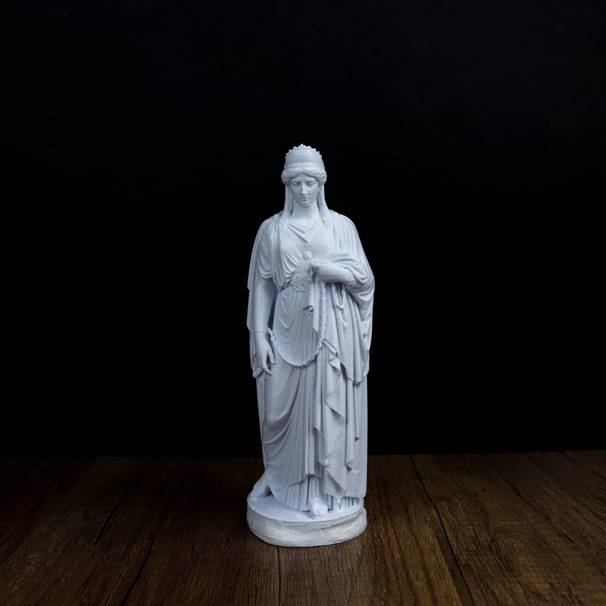A white marble statue of a woman with a flowing robe and headdress stands on a wooden surface against a black background.