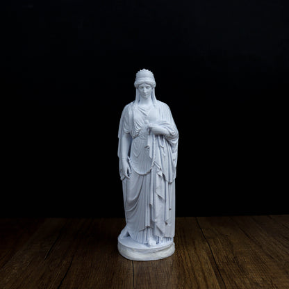 A white marble statue of a woman with a flowing robe and headdress stands on a wooden surface against a black background.