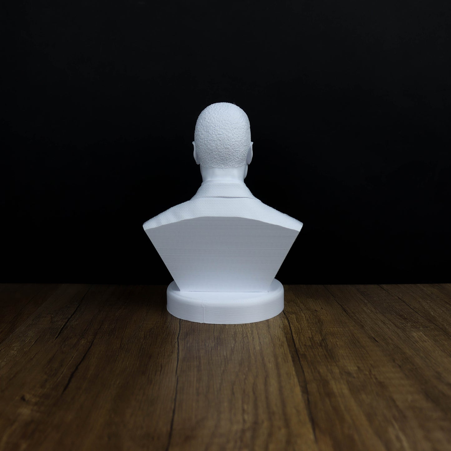 A white bust sculpture is displayed on a wooden surface against a black background.