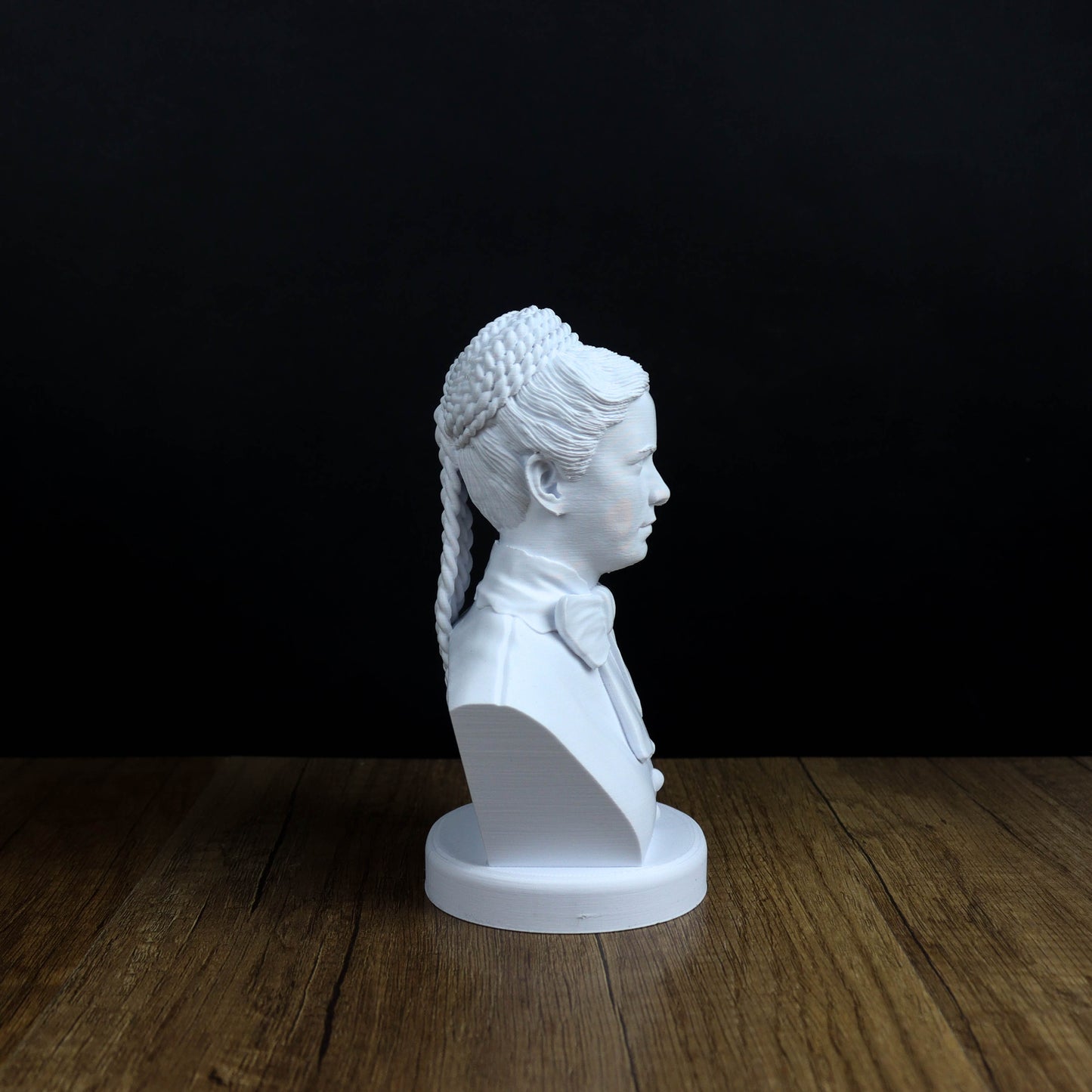A white bust of a person with braided hair is displayed on a wooden surface against a black background.