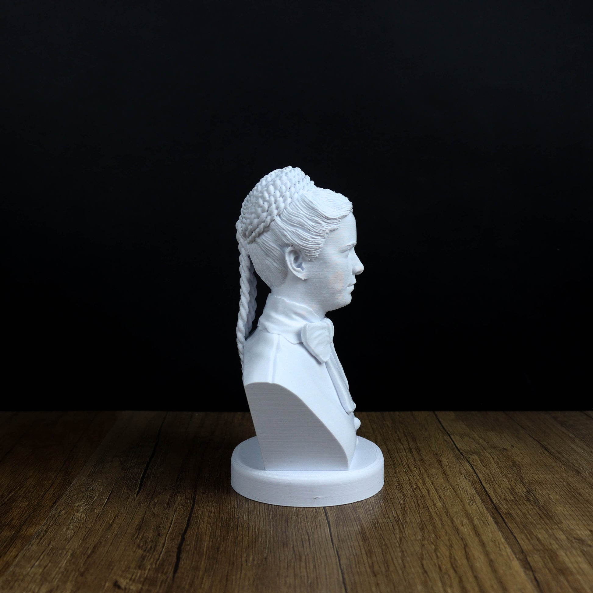 A white bust of a person with braided hair is displayed on a wooden surface against a black background.