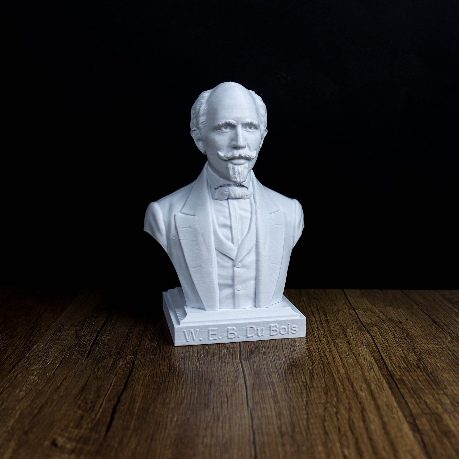 A white bust of a man wearing a suit and tie, sitting on a wooden surface against a dark background.