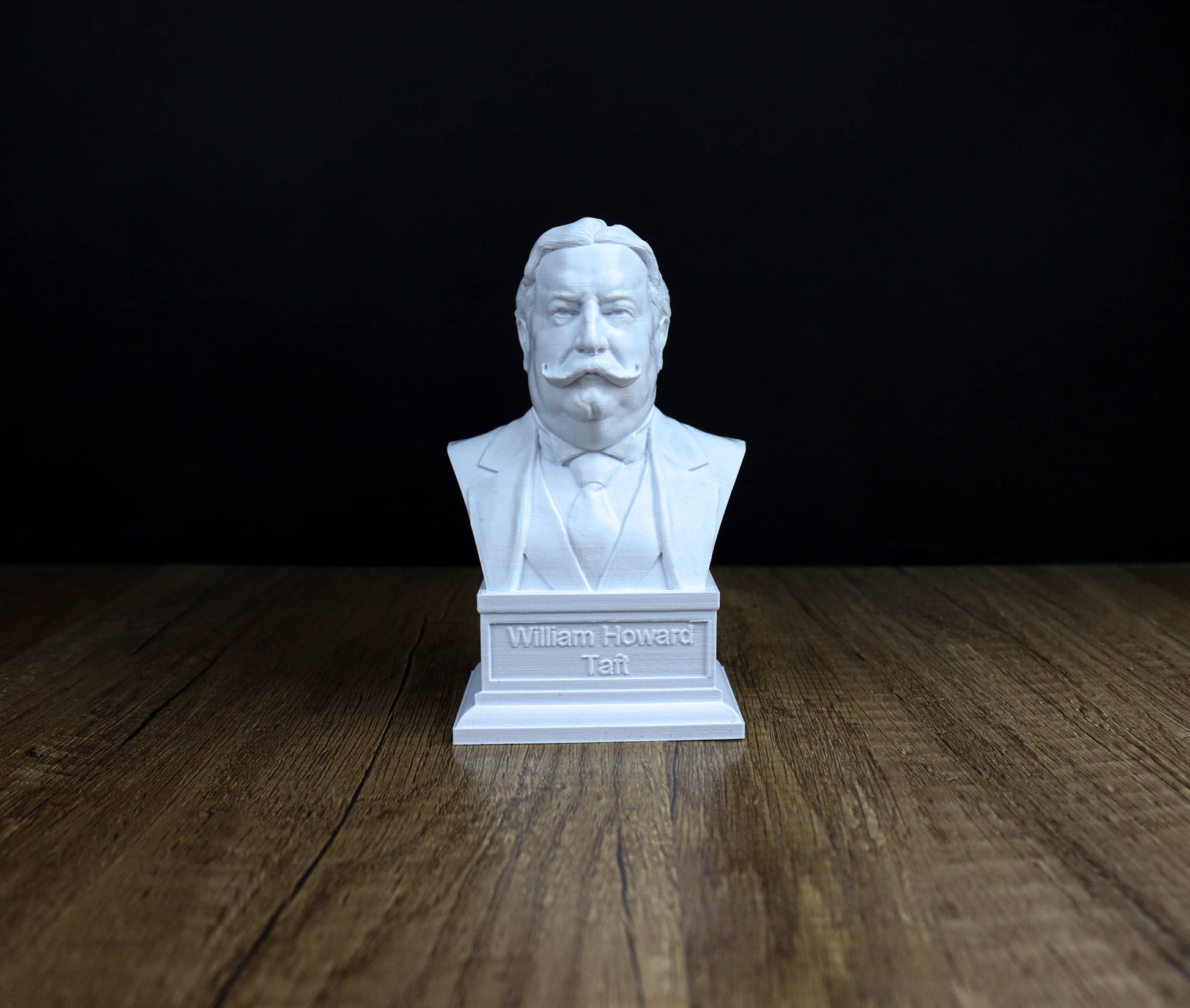 A white bust of a man with a mustache and goatee, sitting on a wooden surface against a black background.
