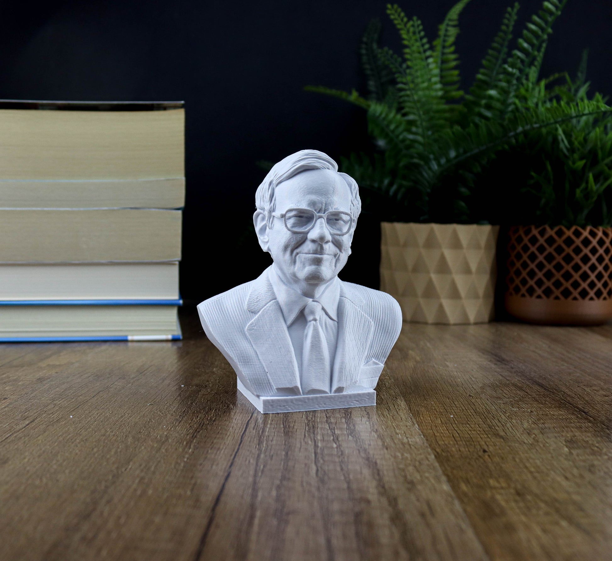 A white bust of a man wearing a suit and tie is displayed on a wooden surface, with a stack of books and a potted plant in the background.