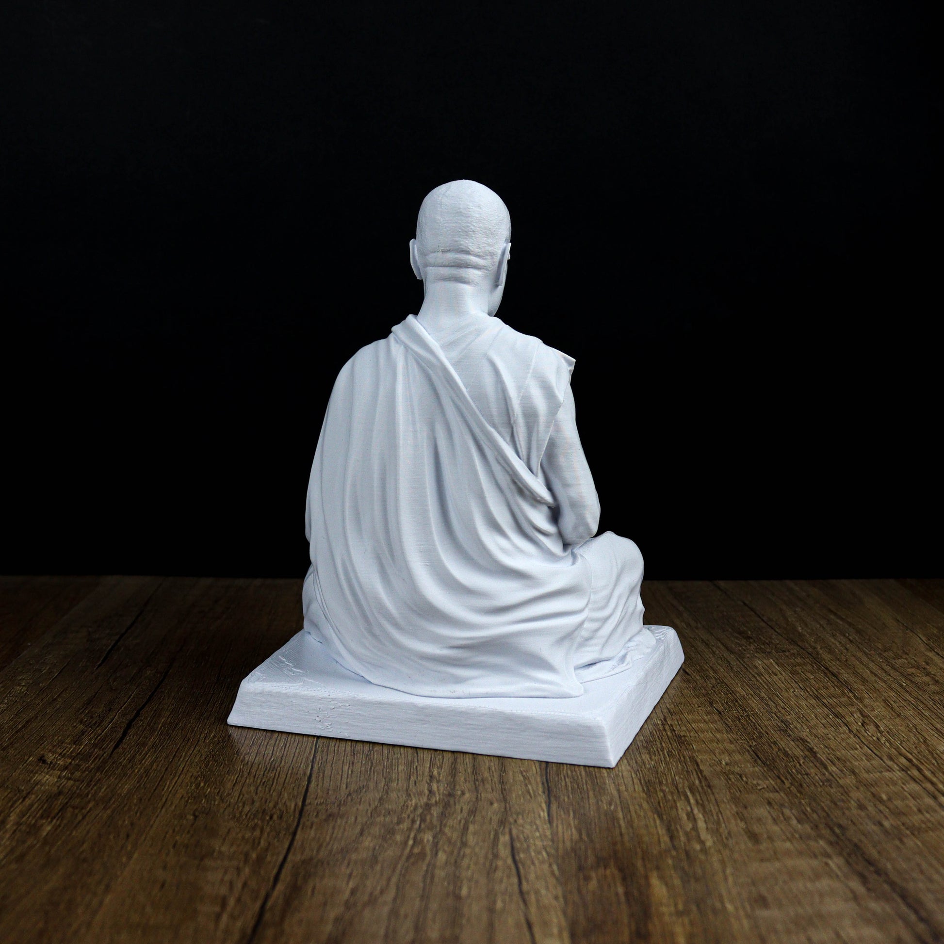 A white statue of a seated Buddha figure is displayed on a wooden surface against a black background.
