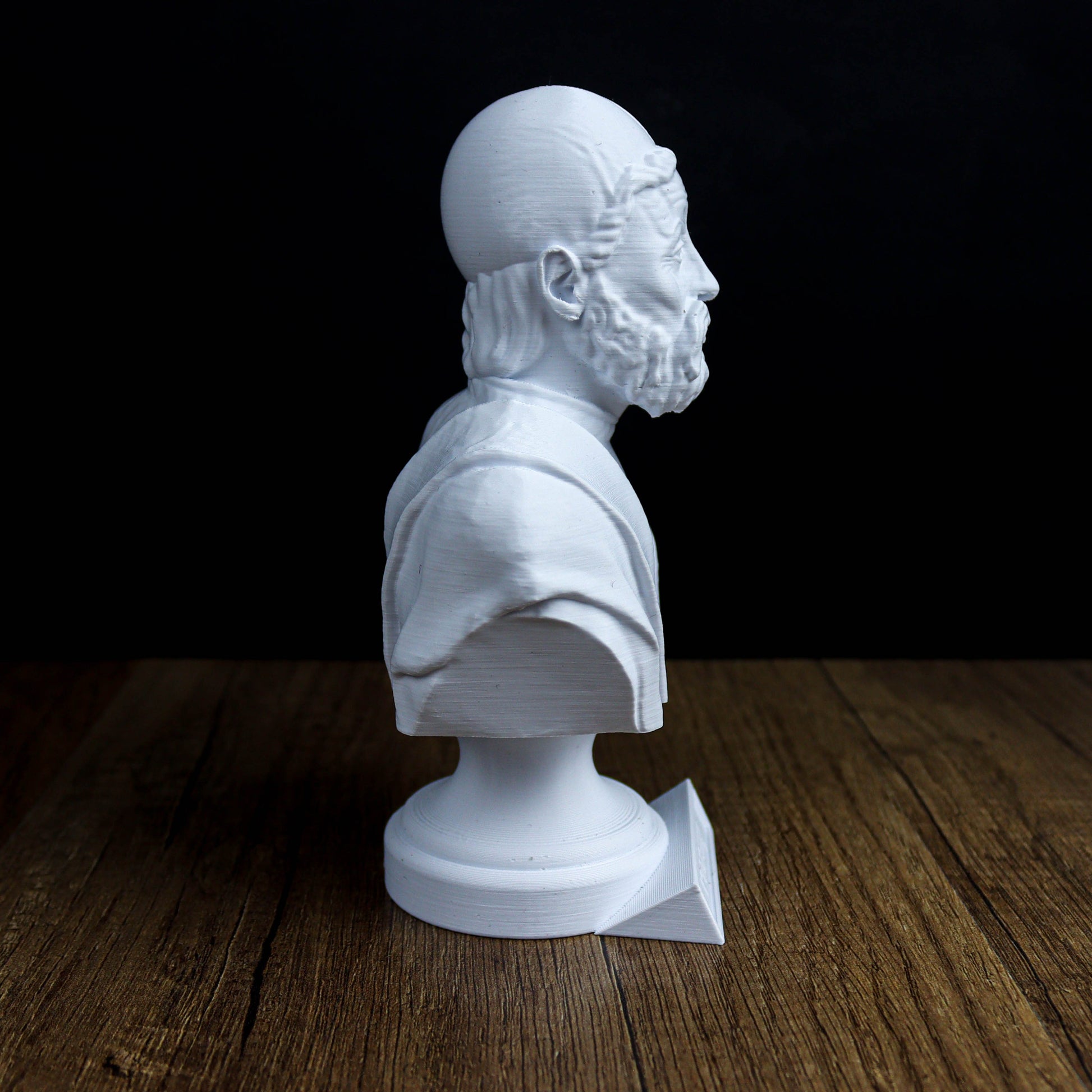 A white bust of a bearded man is displayed on a wooden surface against a dark background.