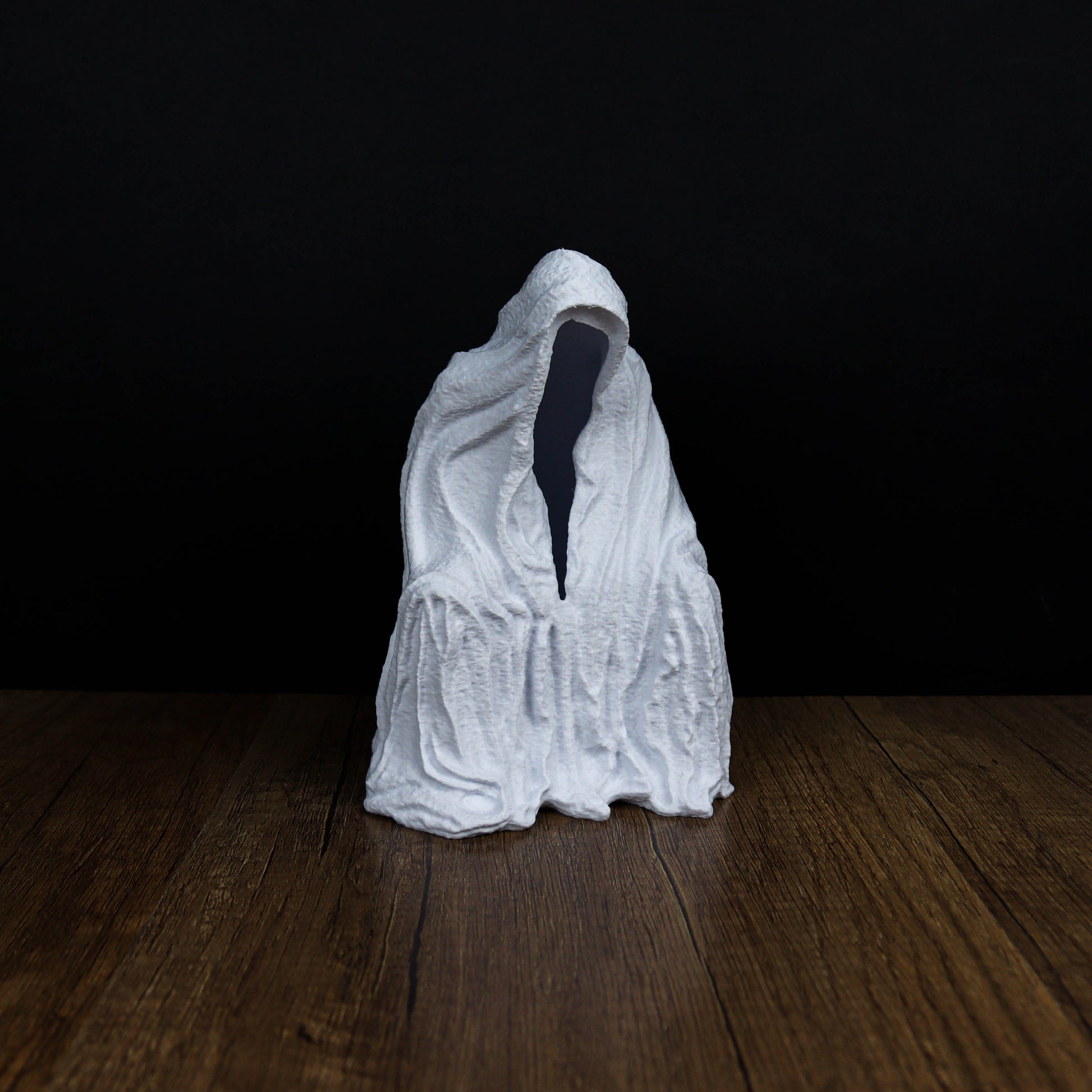 A white, hooded figure sits on a wooden surface against a dark background.
