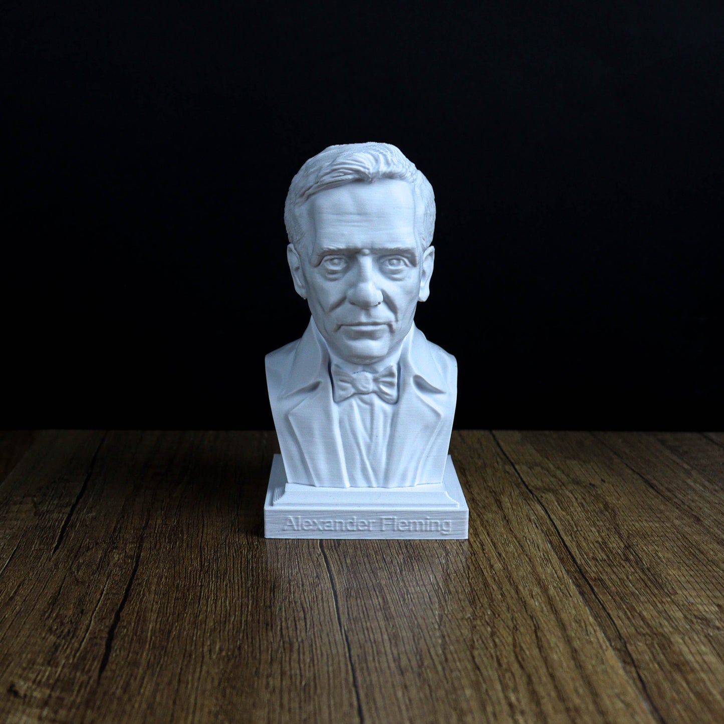 A white bust of a man wearing a suit and bow tie, sitting on a wooden surface against a black background.