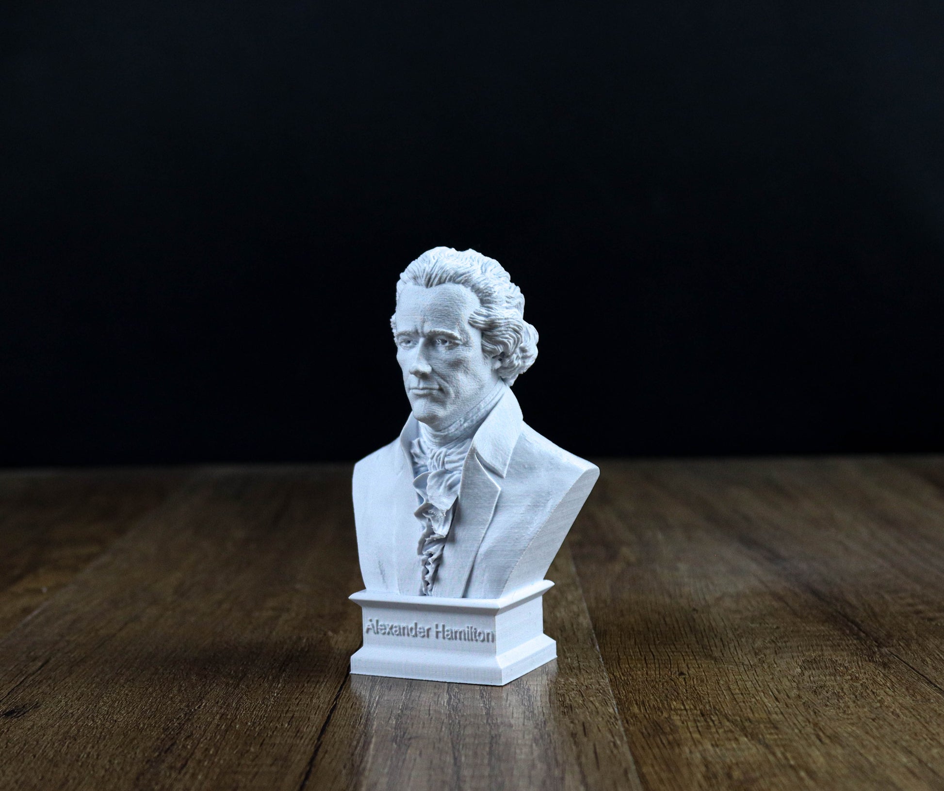 A white bust of a man, likely Alexander Hamilton, is displayed on a wooden surface against a dark background.