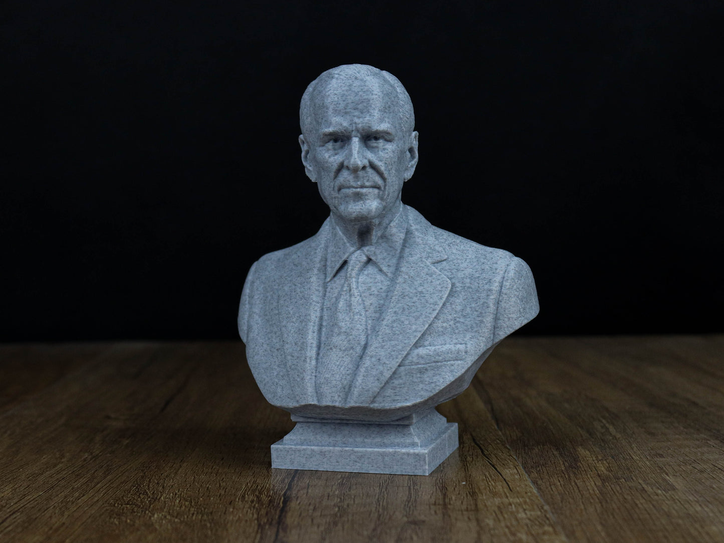A gray stone bust of a man wearing a suit and tie, sitting on a wooden surface against a dark background.