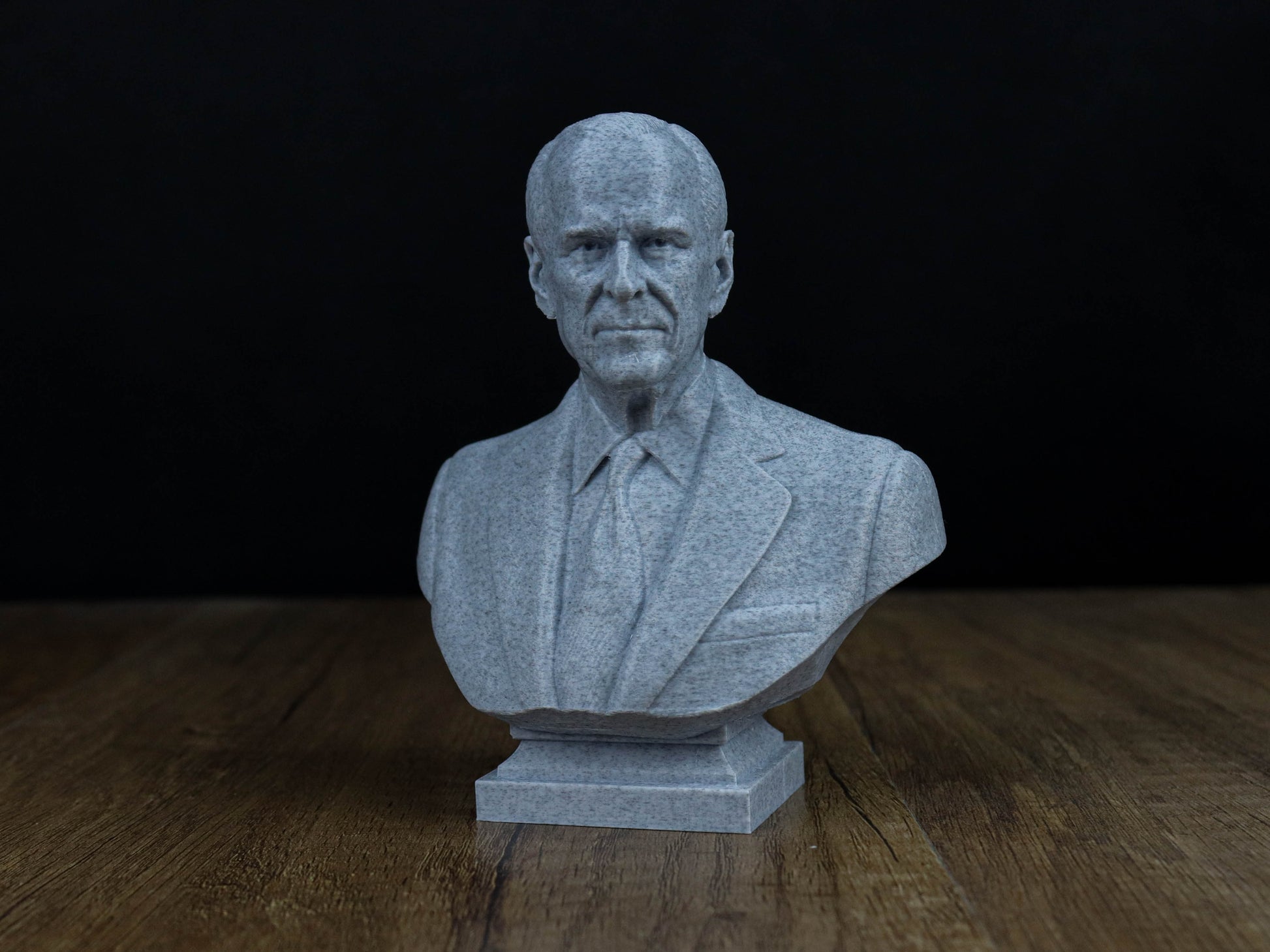A gray stone bust of a man wearing a suit and tie, sitting on a wooden surface against a dark background.