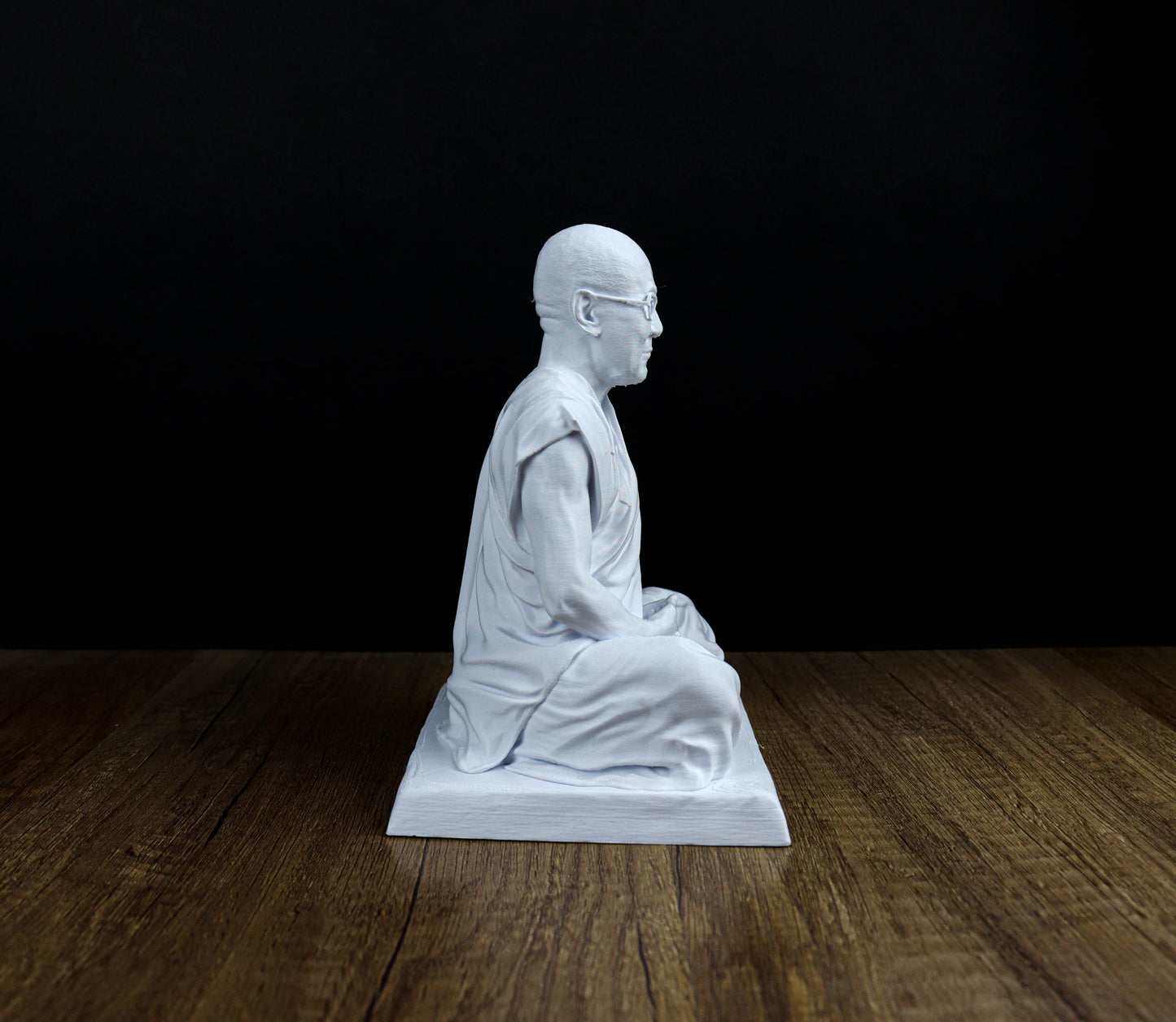 A white statue of a seated Buddha figure is displayed on a wooden surface against a black background.