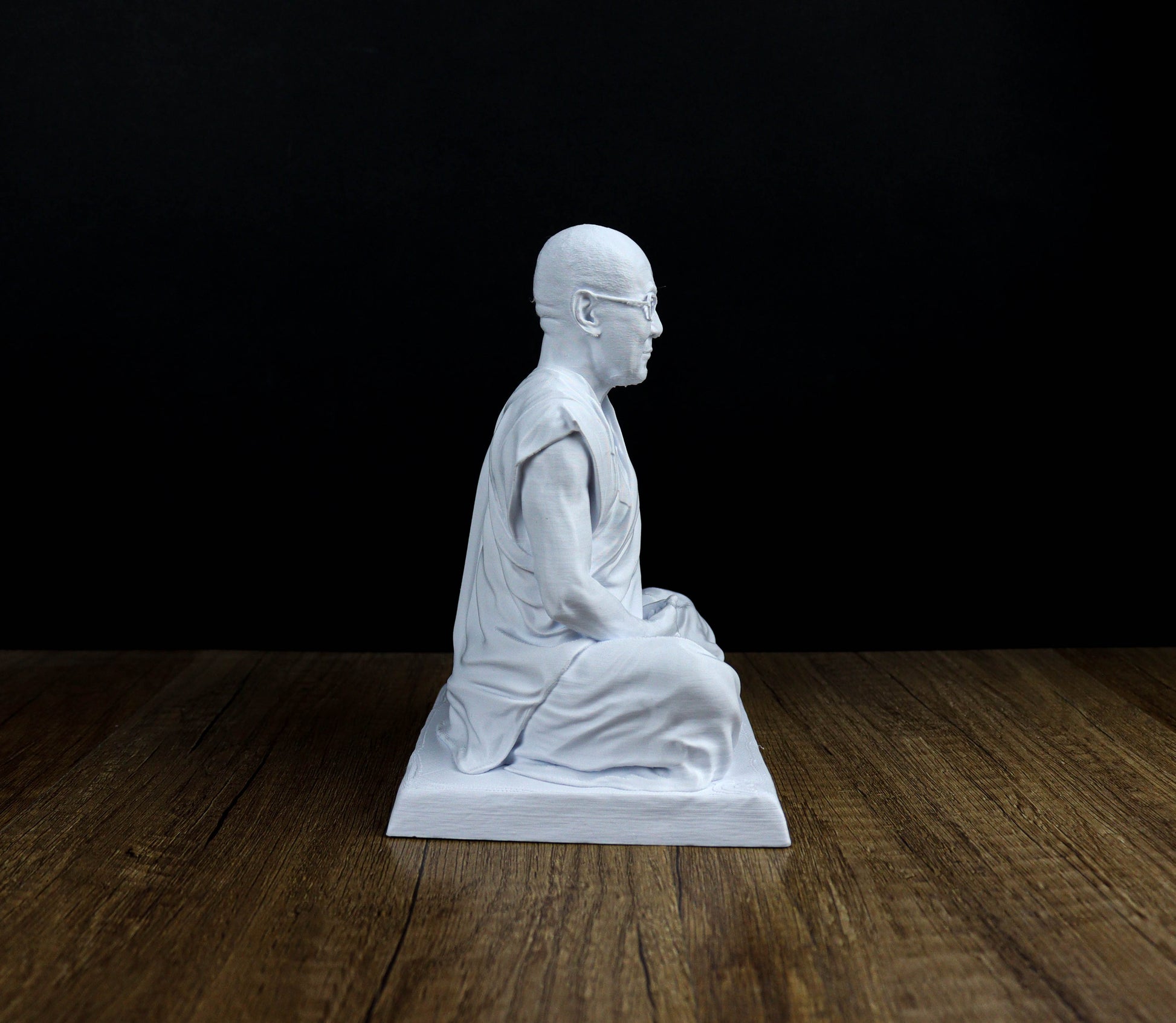 A white statue of a seated Buddha figure is displayed on a wooden surface against a black background.