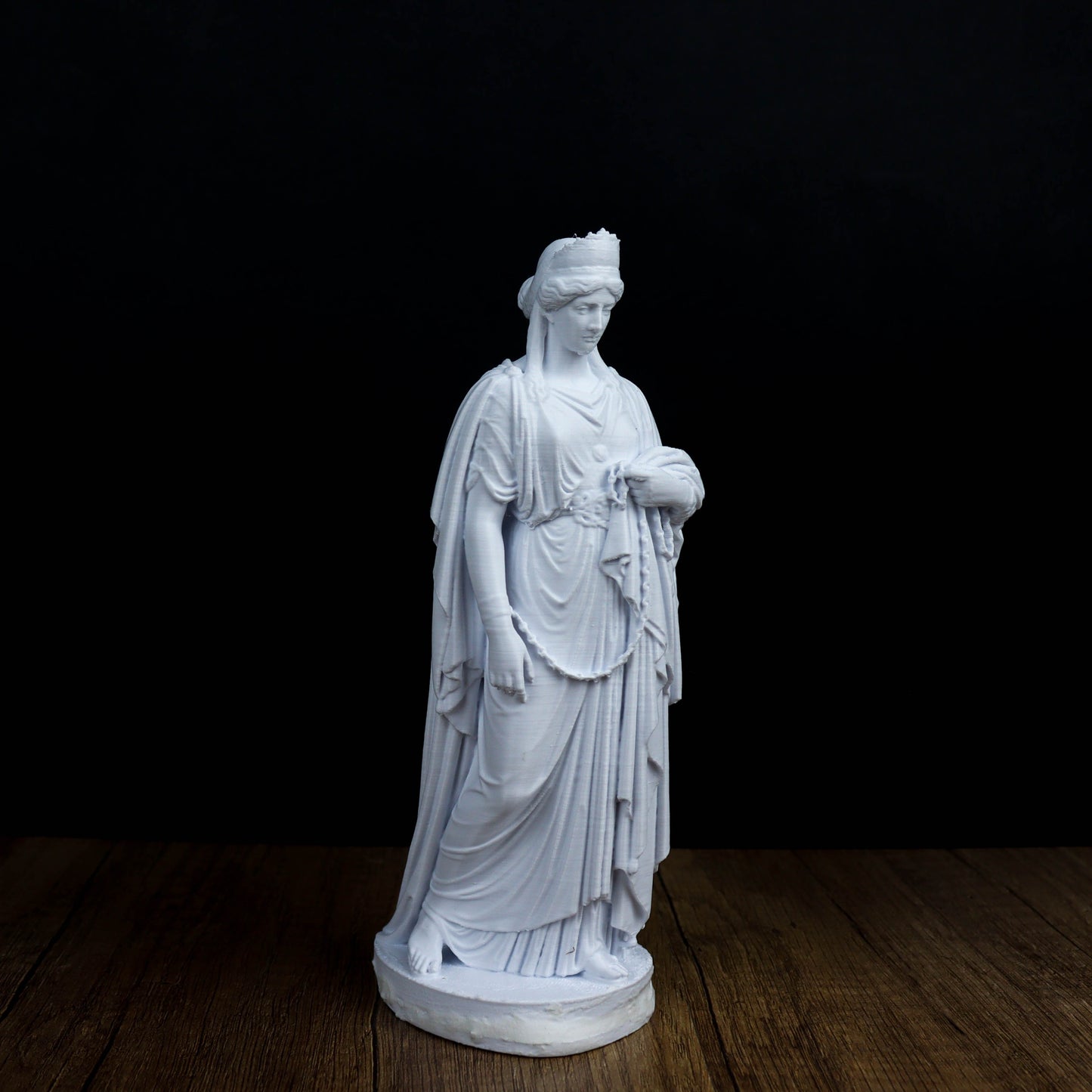 A white marble statue of a woman with a flowing robe and headdress stands on a wooden surface against a black background.