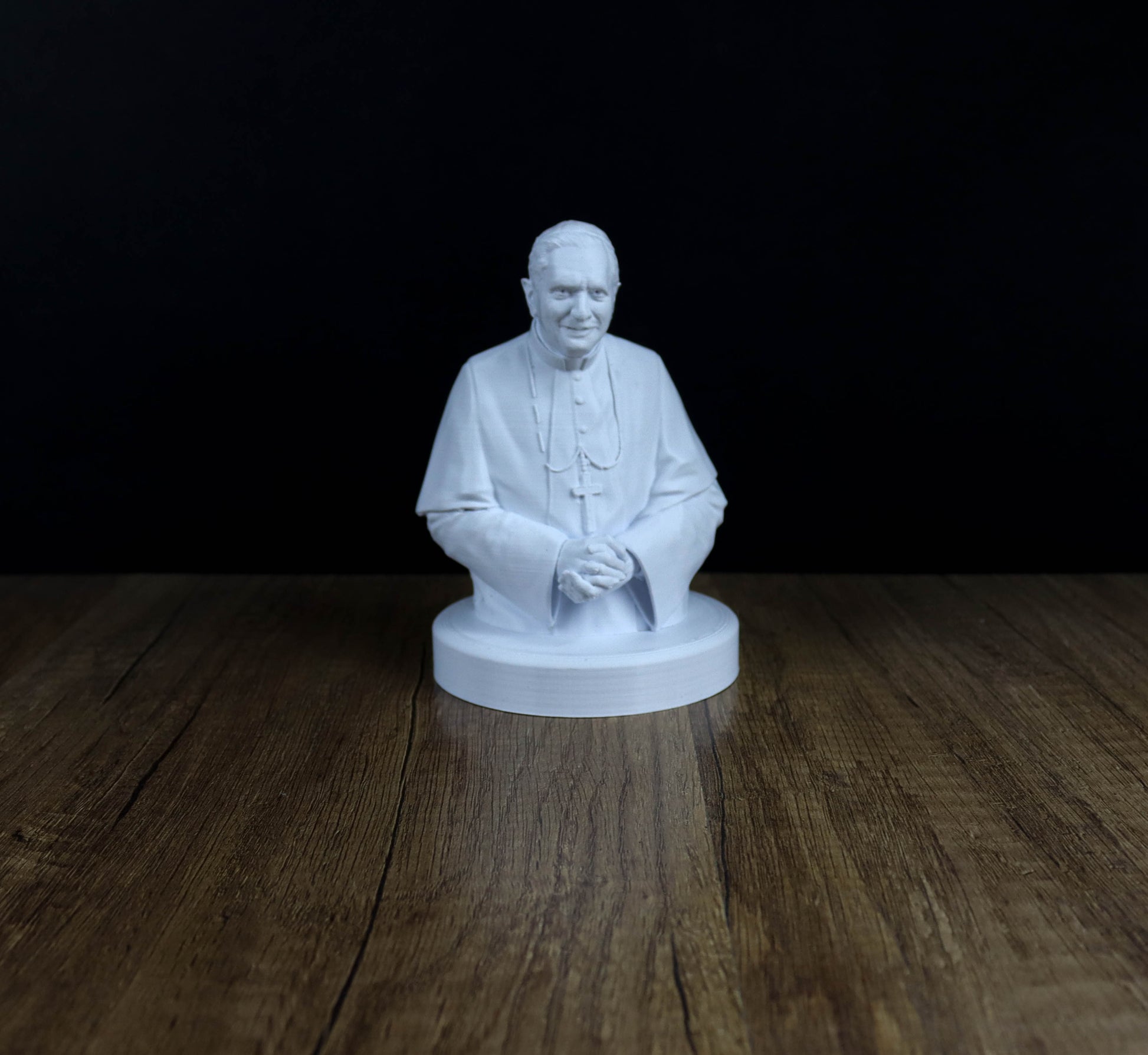 A white bust of a person, possibly a religious figure, sitting on a wooden surface against a dark background.