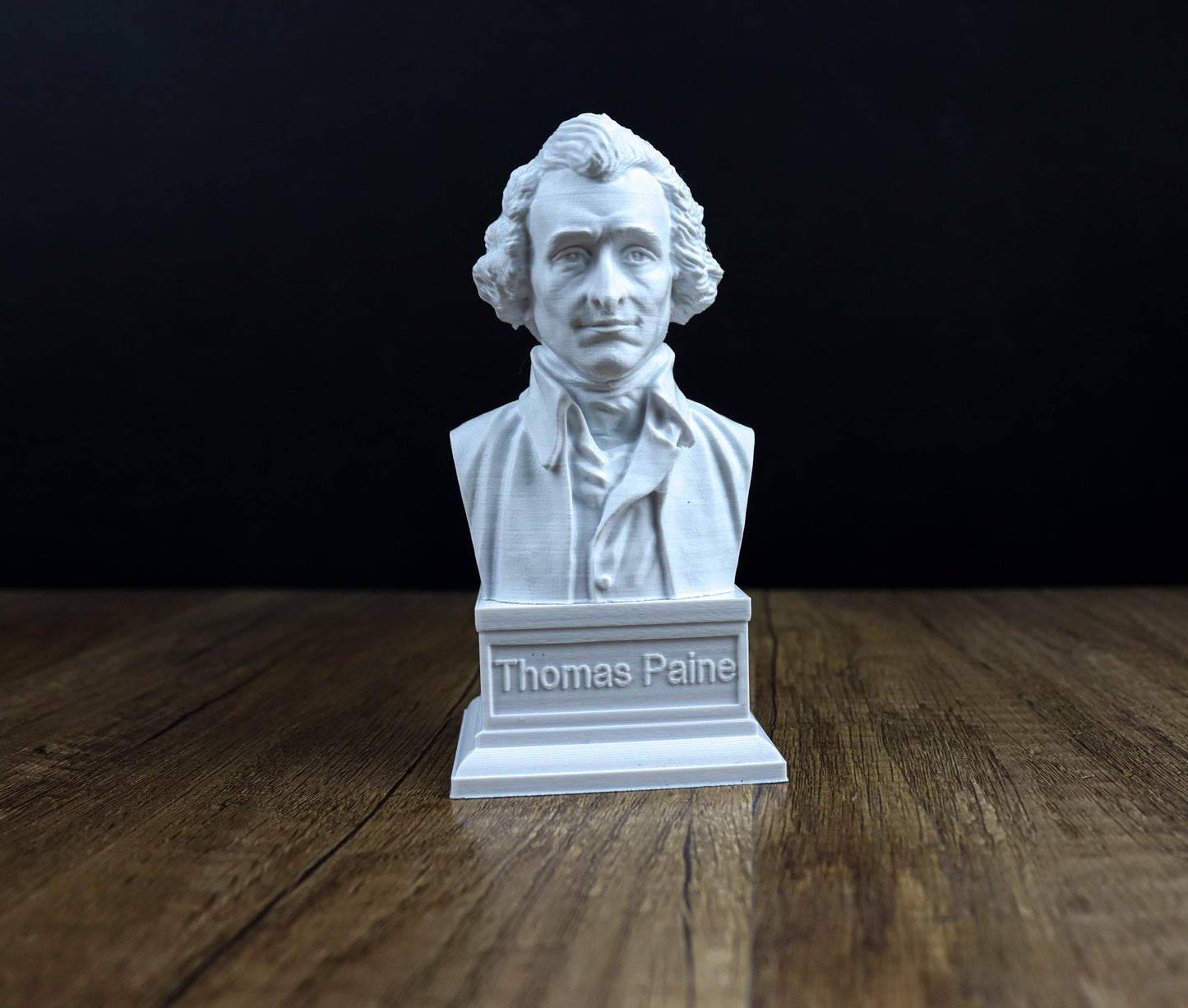 A white bust of a man, likely Thomas Paine, is displayed on a wooden surface against a dark background.
