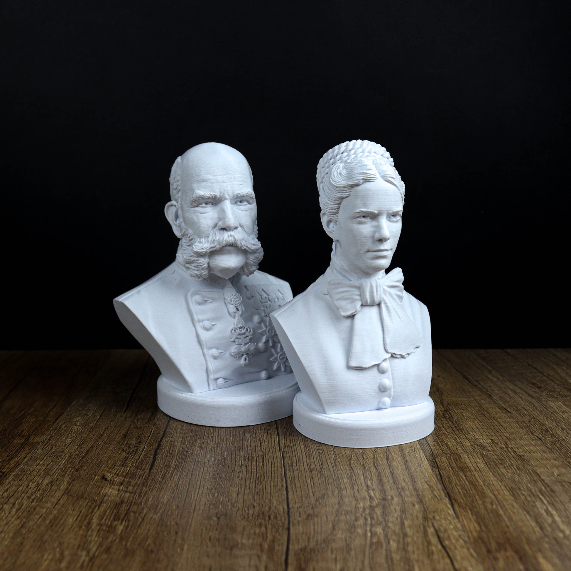 Two white busts of men, one with a beard and the other with a bow tie, are displayed on a wooden surface against a black background.