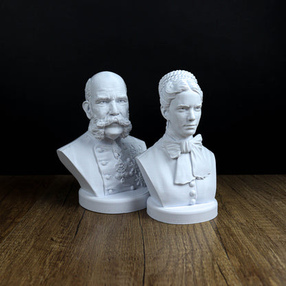 Two white busts of men, one with a beard and the other with a bow tie, are displayed on a wooden surface against a black background.