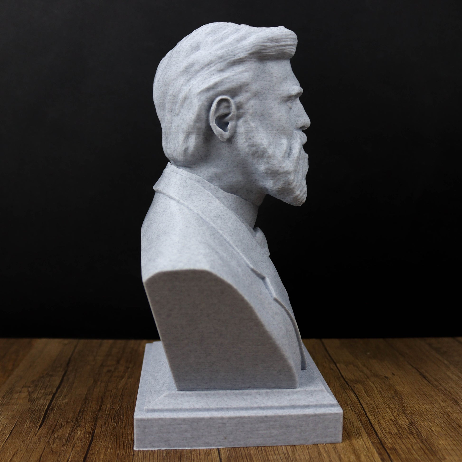 A gray bust sculpture of a man with a beard is displayed on a wooden surface against a black background.