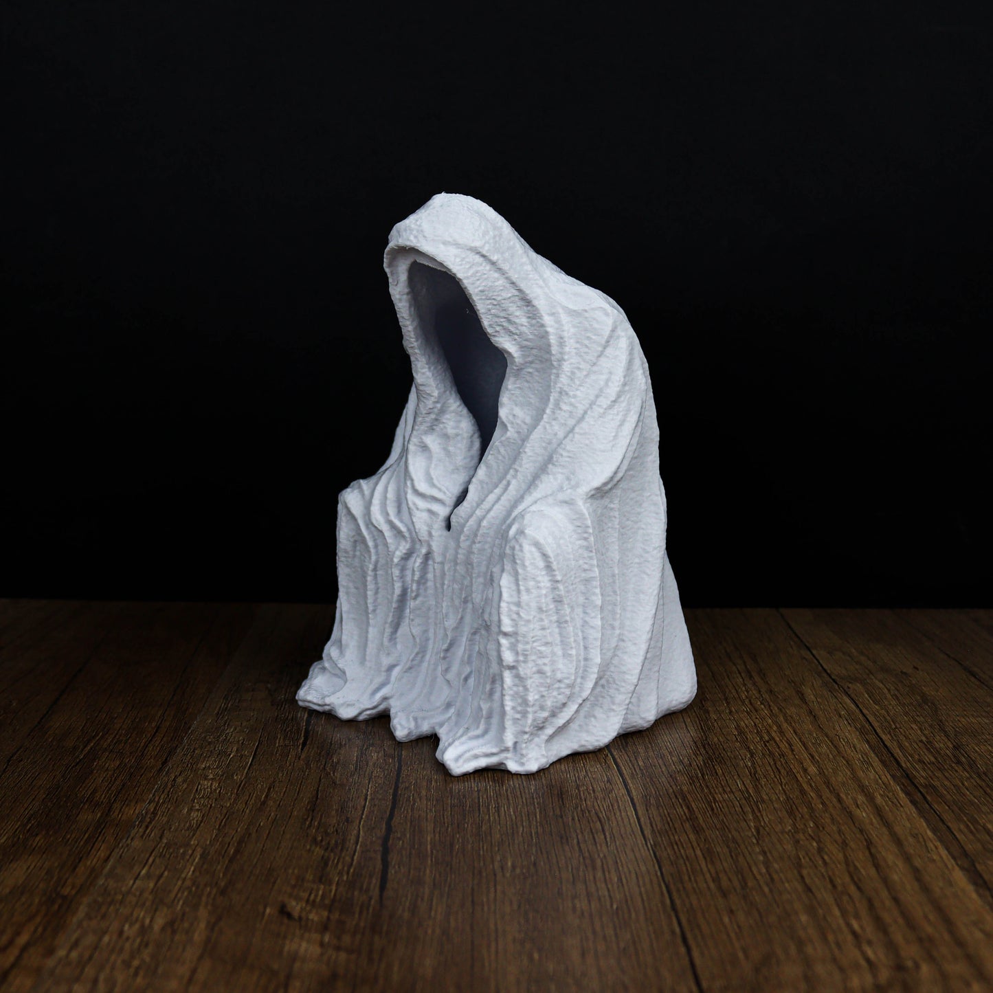 A white, hooded figure sits on a wooden surface against a dark background.