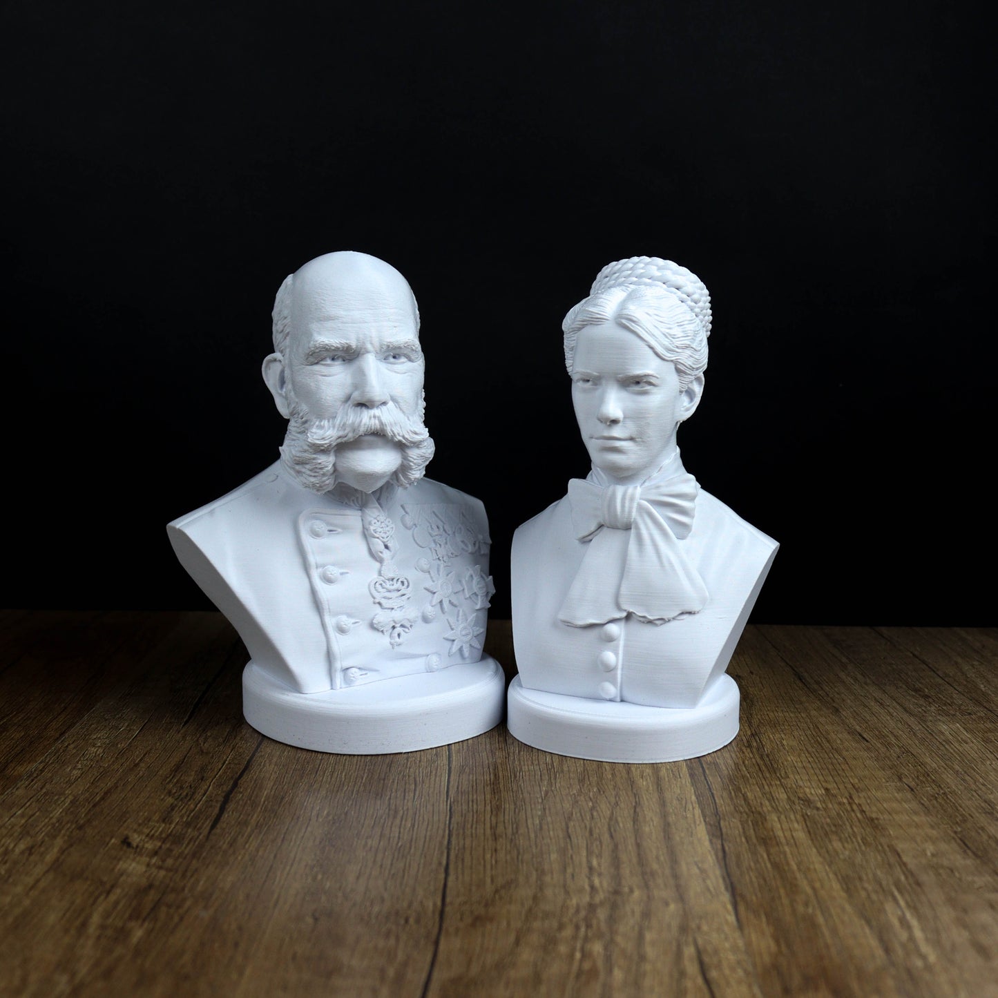 Two white busts of a man and a woman, both with distinct facial features and attire, are displayed on a wooden surface against a black background.
