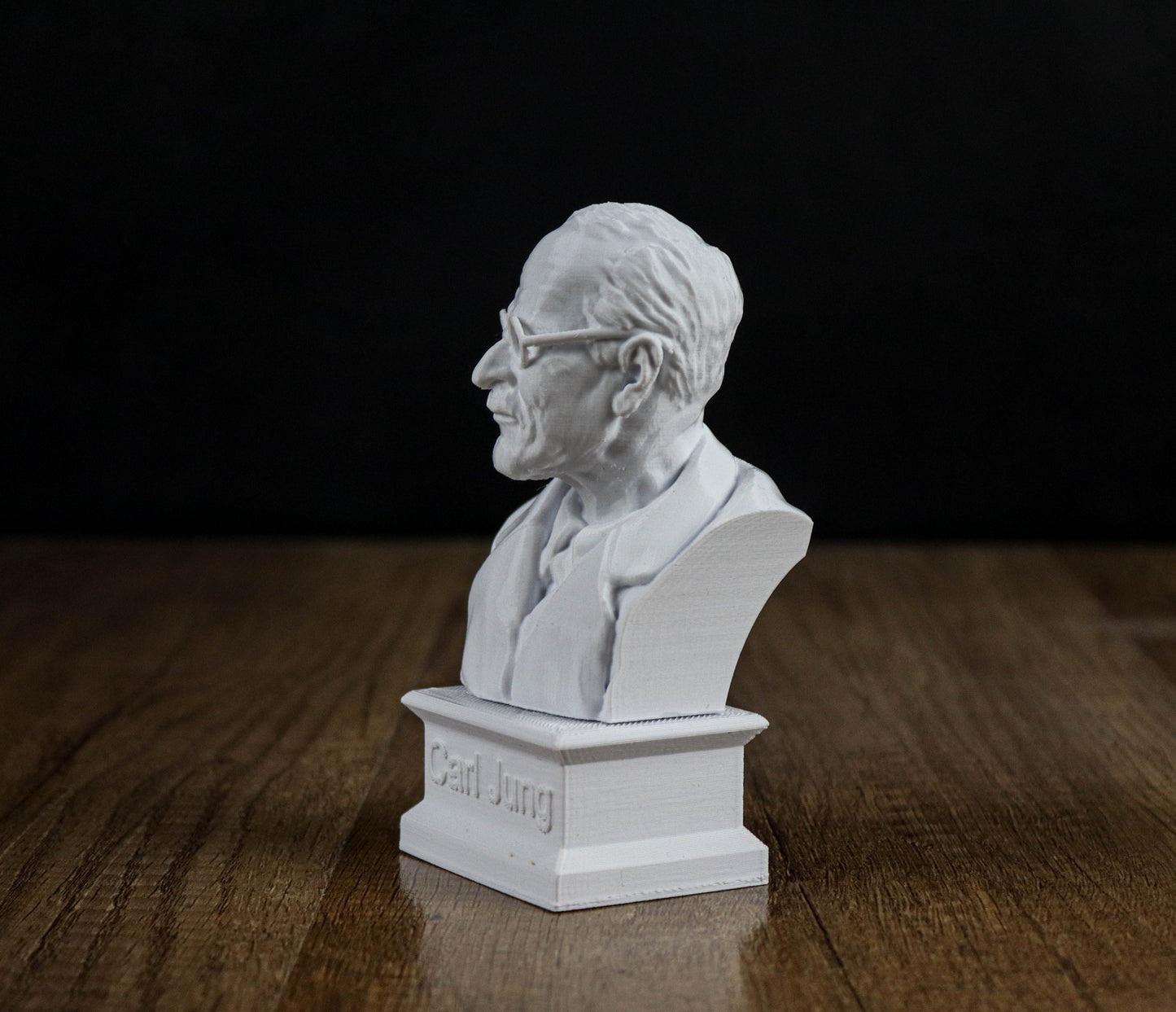 A white bust of a man wearing glasses is displayed on a wooden surface against a black background.