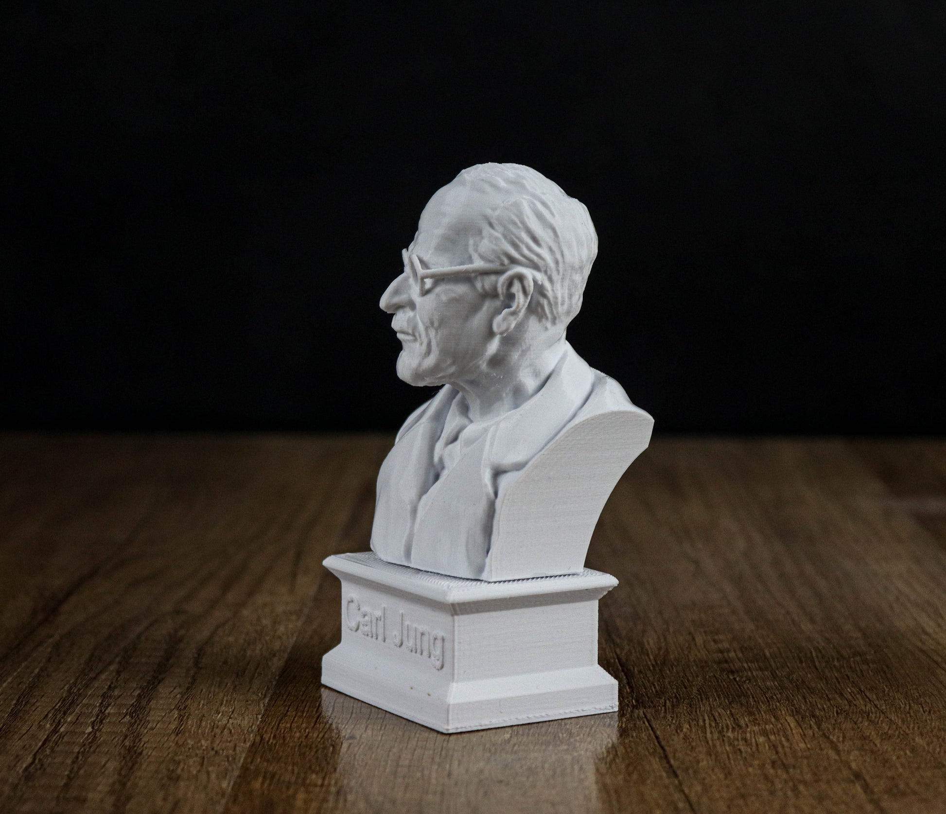 A white bust of a man wearing glasses is displayed on a wooden surface against a black background.