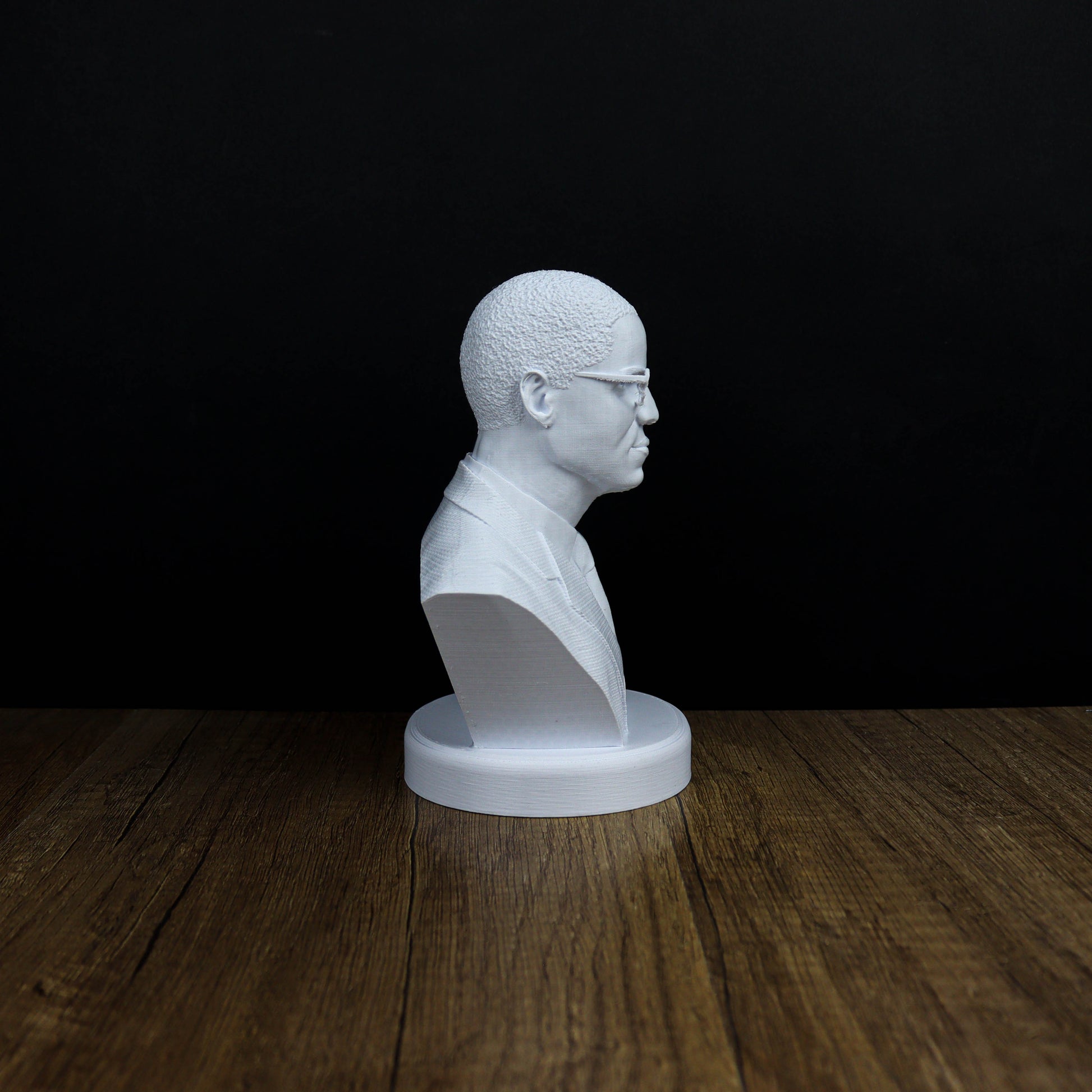 A white bust of a person wearing glasses is displayed on a wooden surface against a black background.