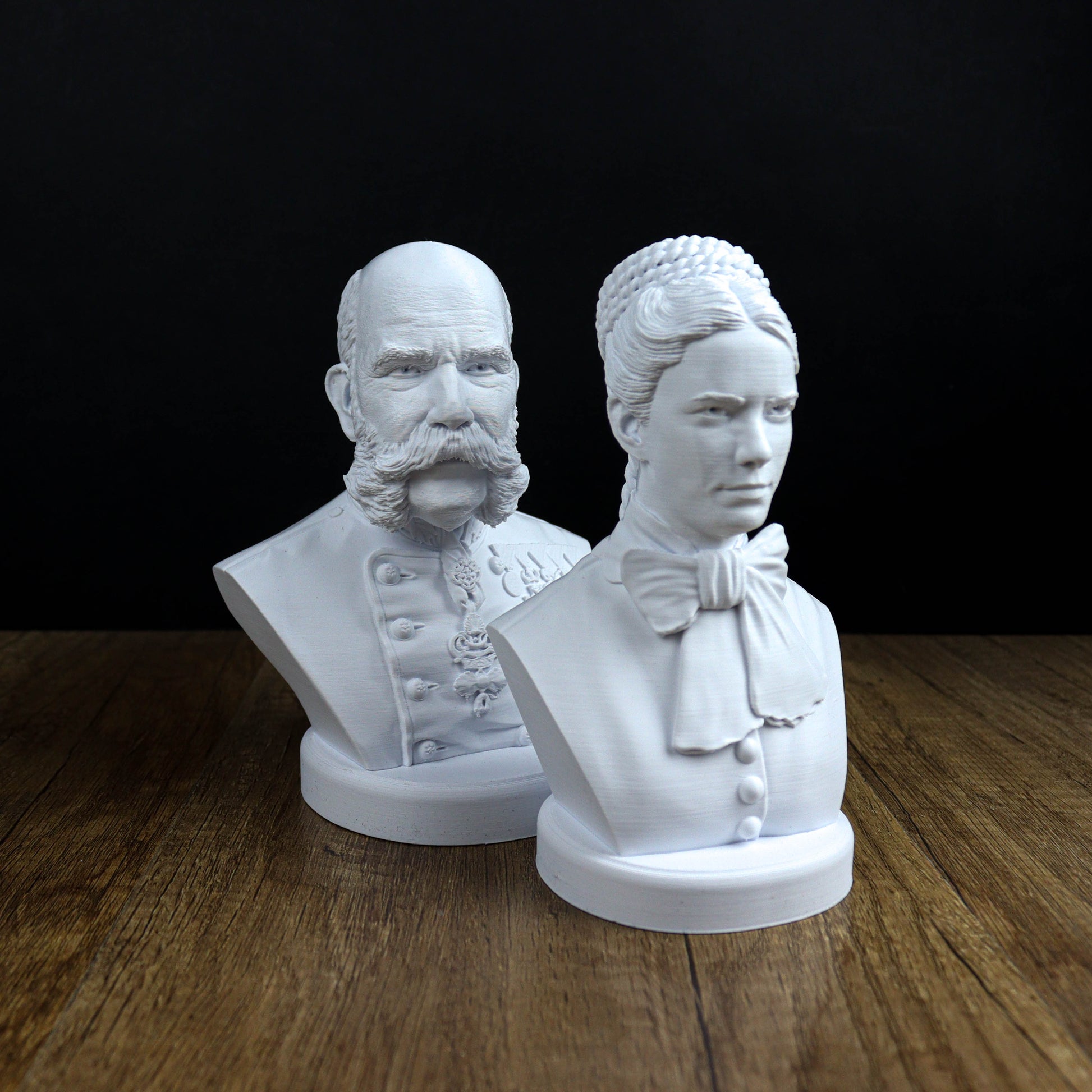 Two white busts of a man and a woman, both with distinct hairstyles, are displayed on a wooden surface against a black background.