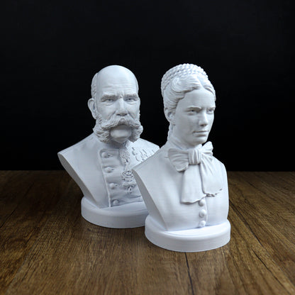 Two white busts of a man and a woman, both with distinct hairstyles, are displayed on a wooden surface against a black background.