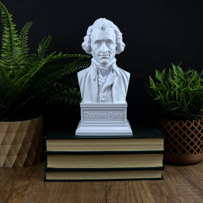 A white bust of Thomas Paine is displayed on top of three books, surrounded by potted plants.