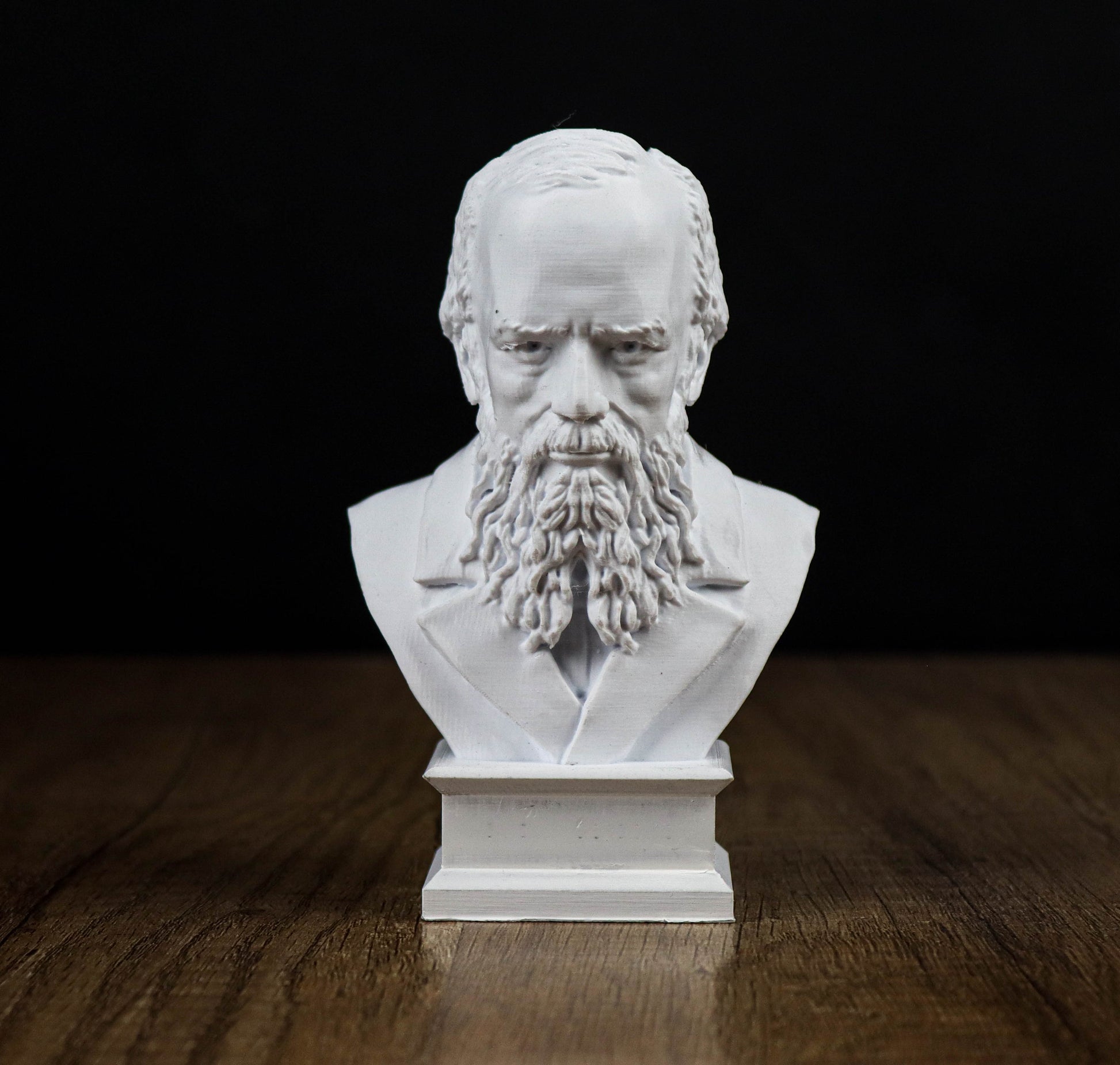 A white bust of a bearded man with a serious expression, sitting on a wooden surface against a black background.