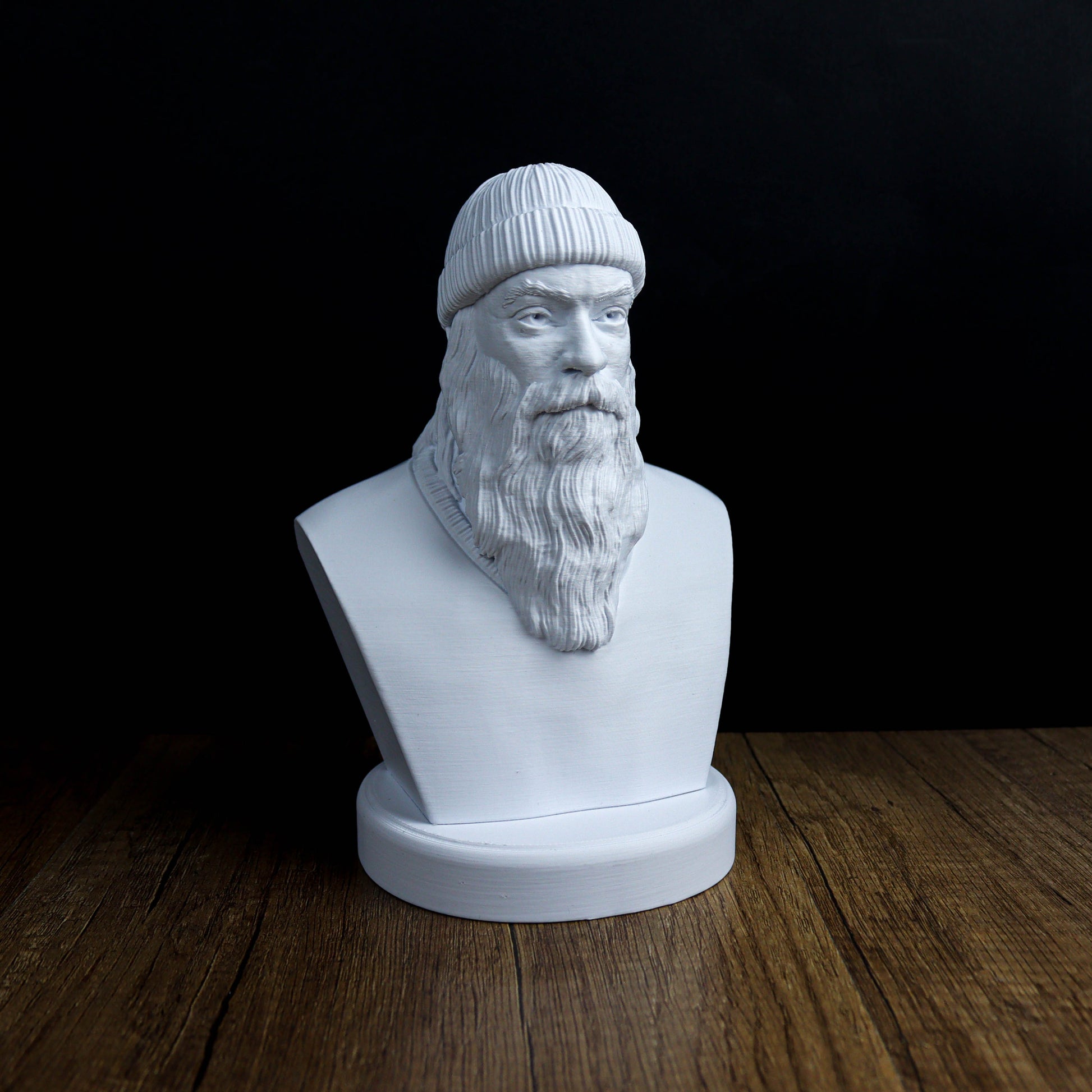 A white bust of a bearded man wearing a hat, sitting on a wooden surface against a black background.