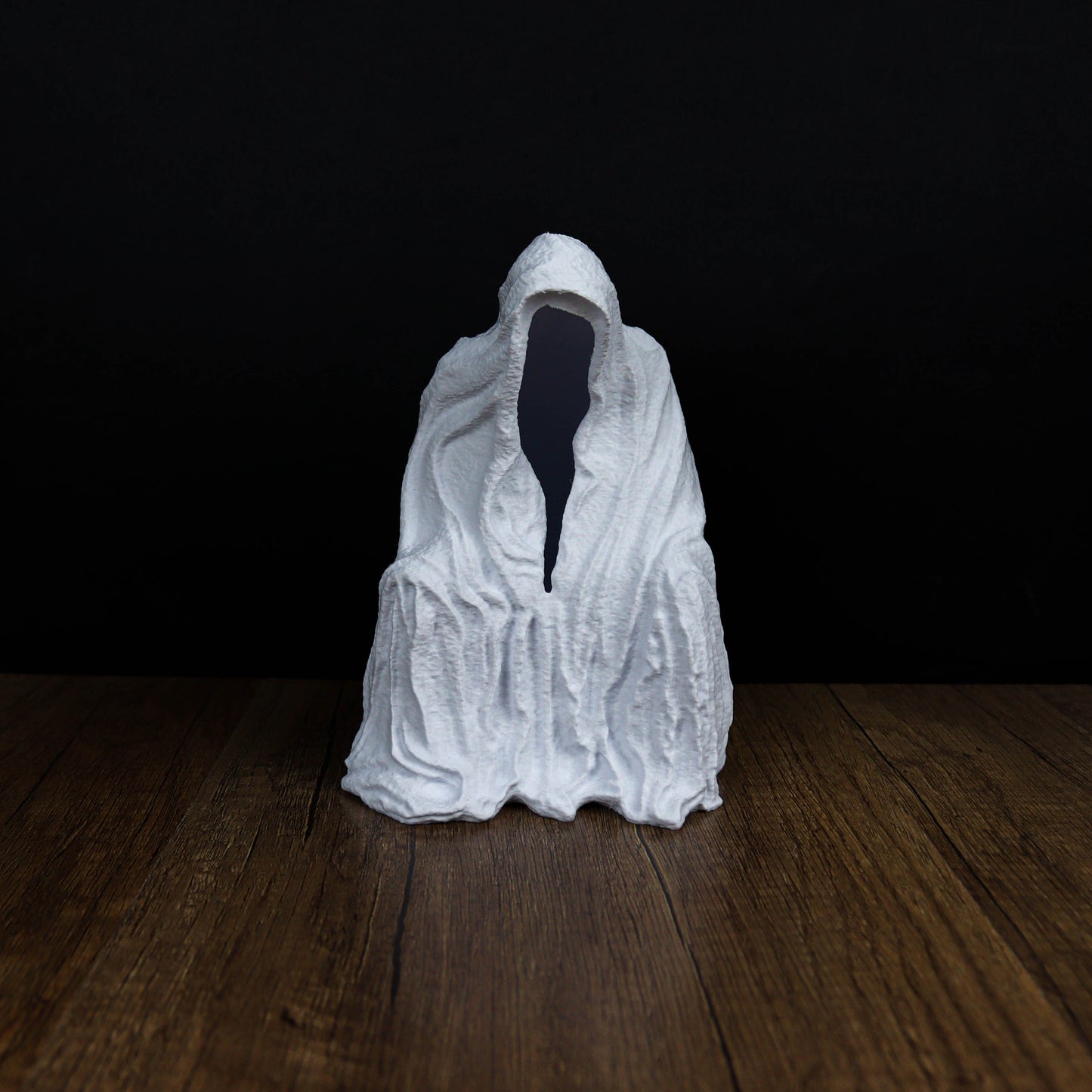 A white, hooded figure sits on a wooden surface against a dark background.