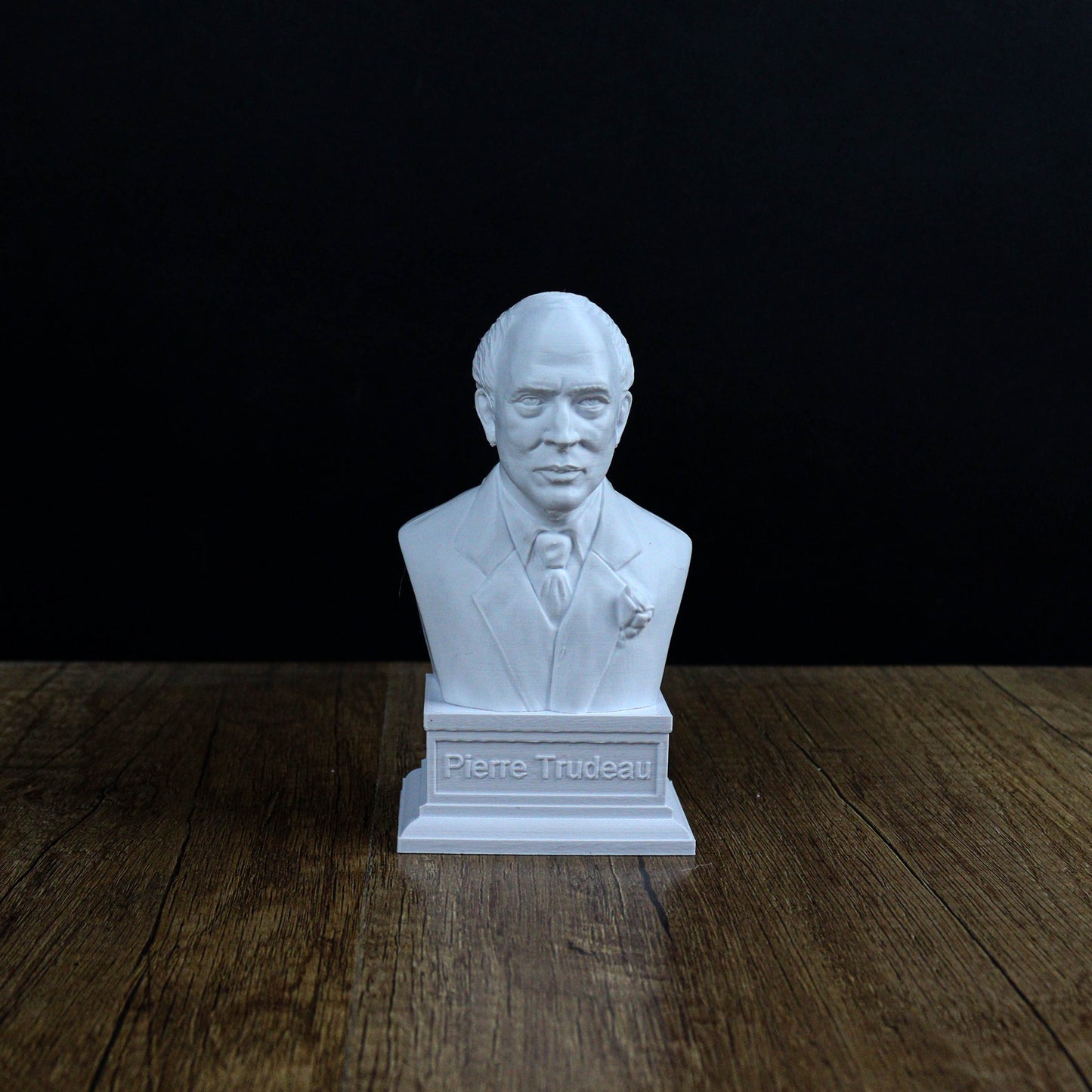 A white bust of a man wearing a suit and tie, with the name "Pierre Trudeau" displayed below it, is sitting on a wooden surface against a dark background.