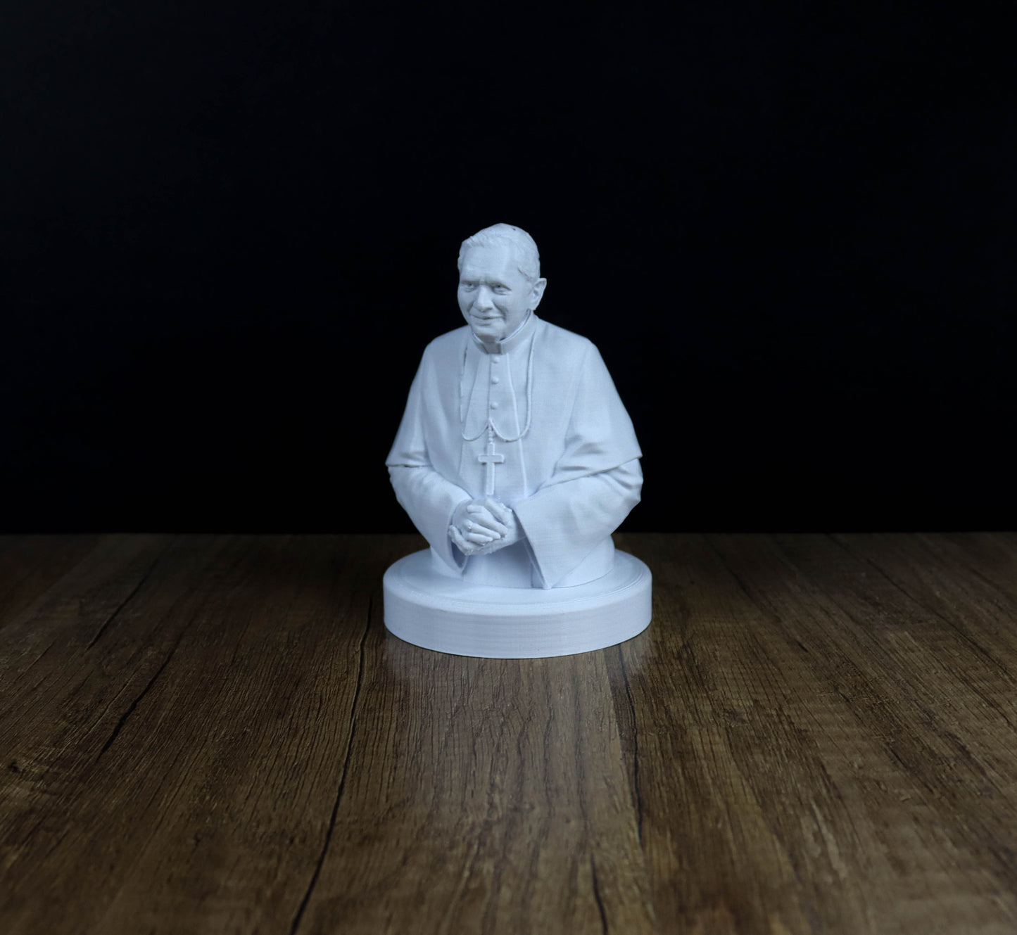 A white 3D model of a person, possibly a religious figure, sitting on a wooden surface against a dark background.
