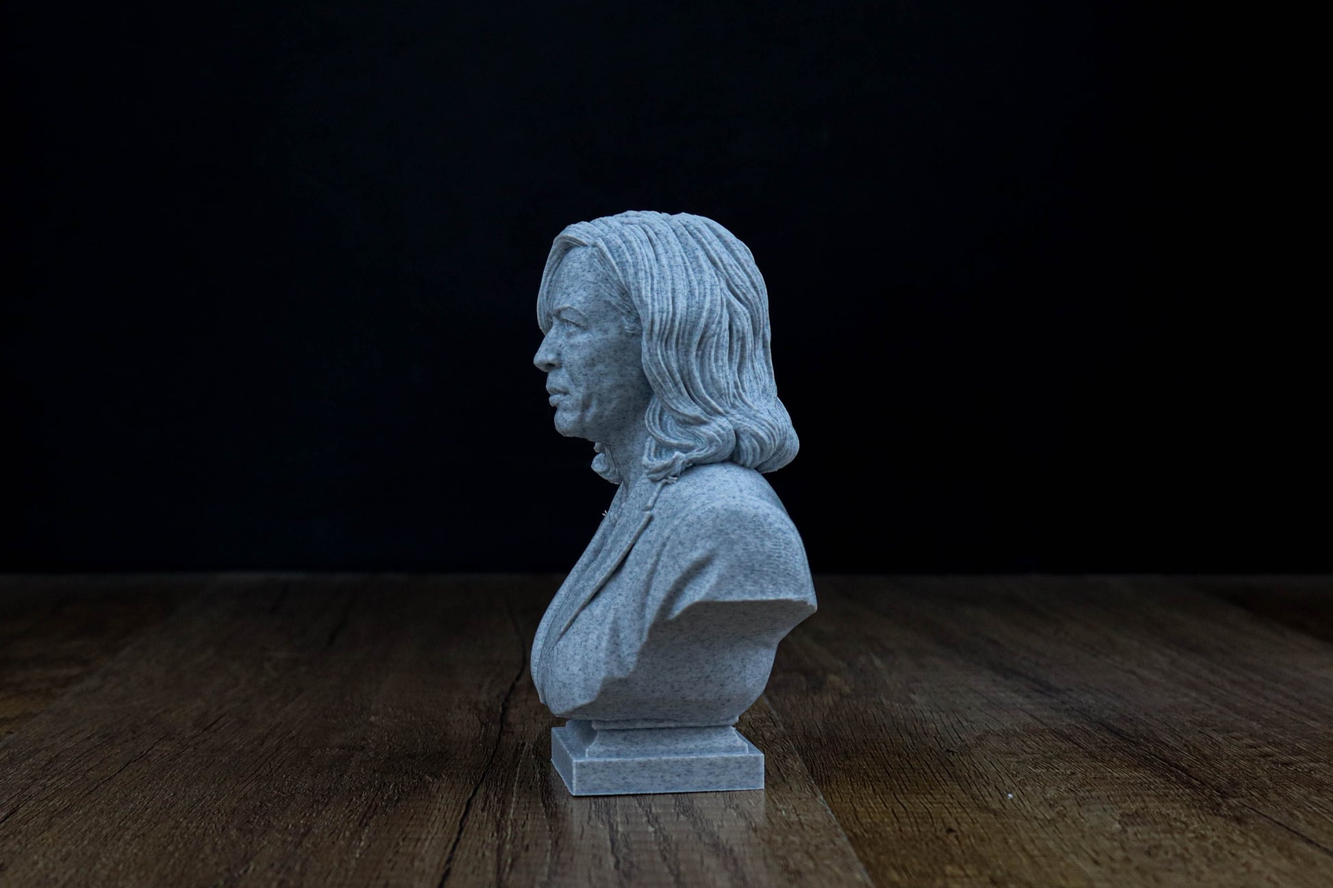 A small gray stone bust of a person is sitting on a wooden surface against a dark background.