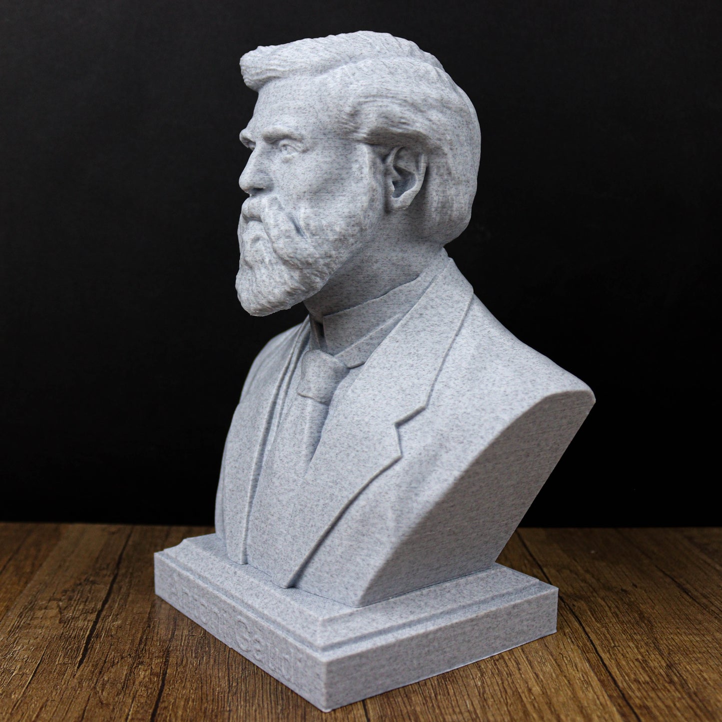 A gray stone bust of a man with a beard, wearing a suit and tie, sitting on a wooden surface.
