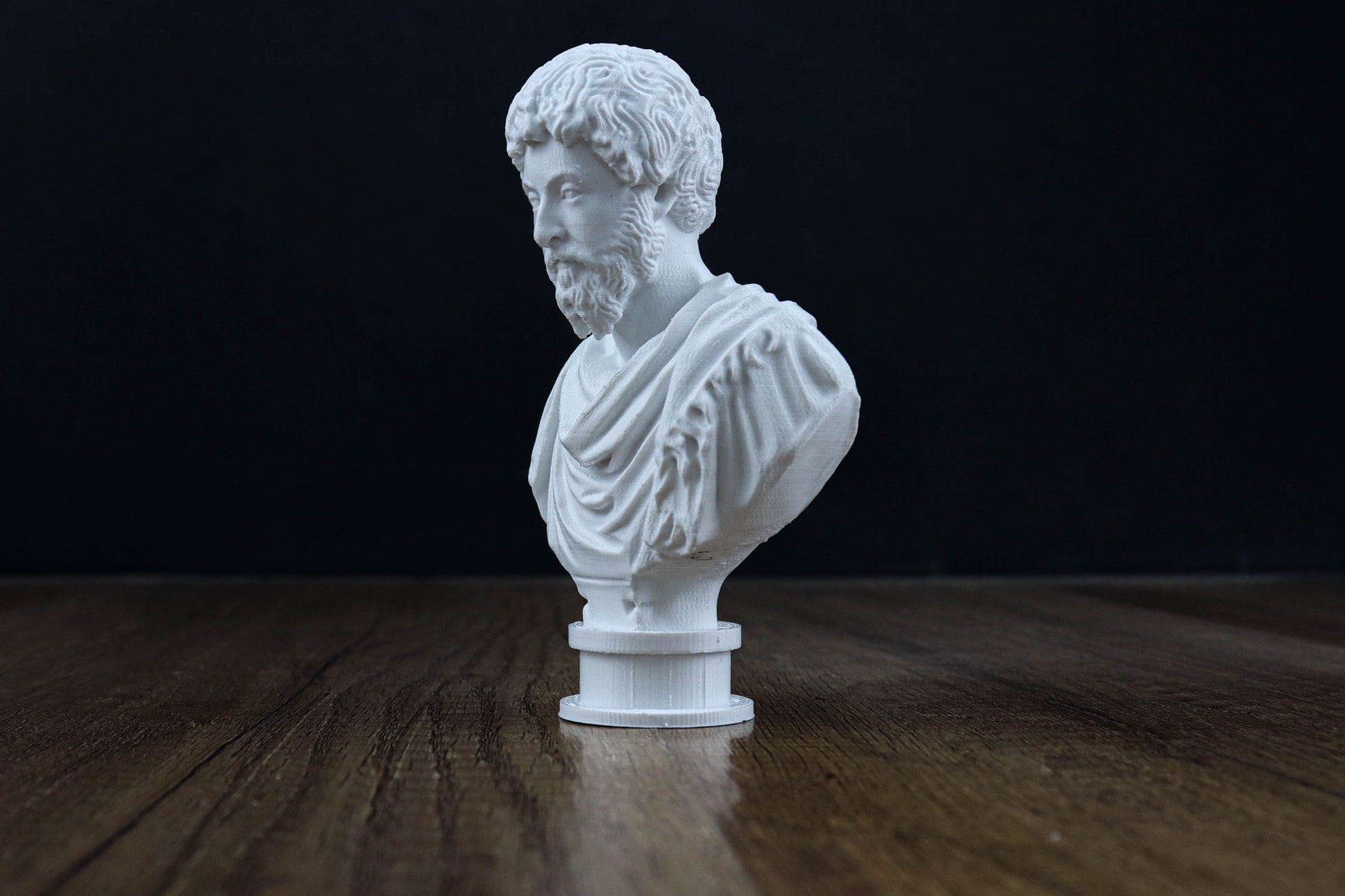 A white bust of a bearded man is displayed on a wooden surface against a dark background.